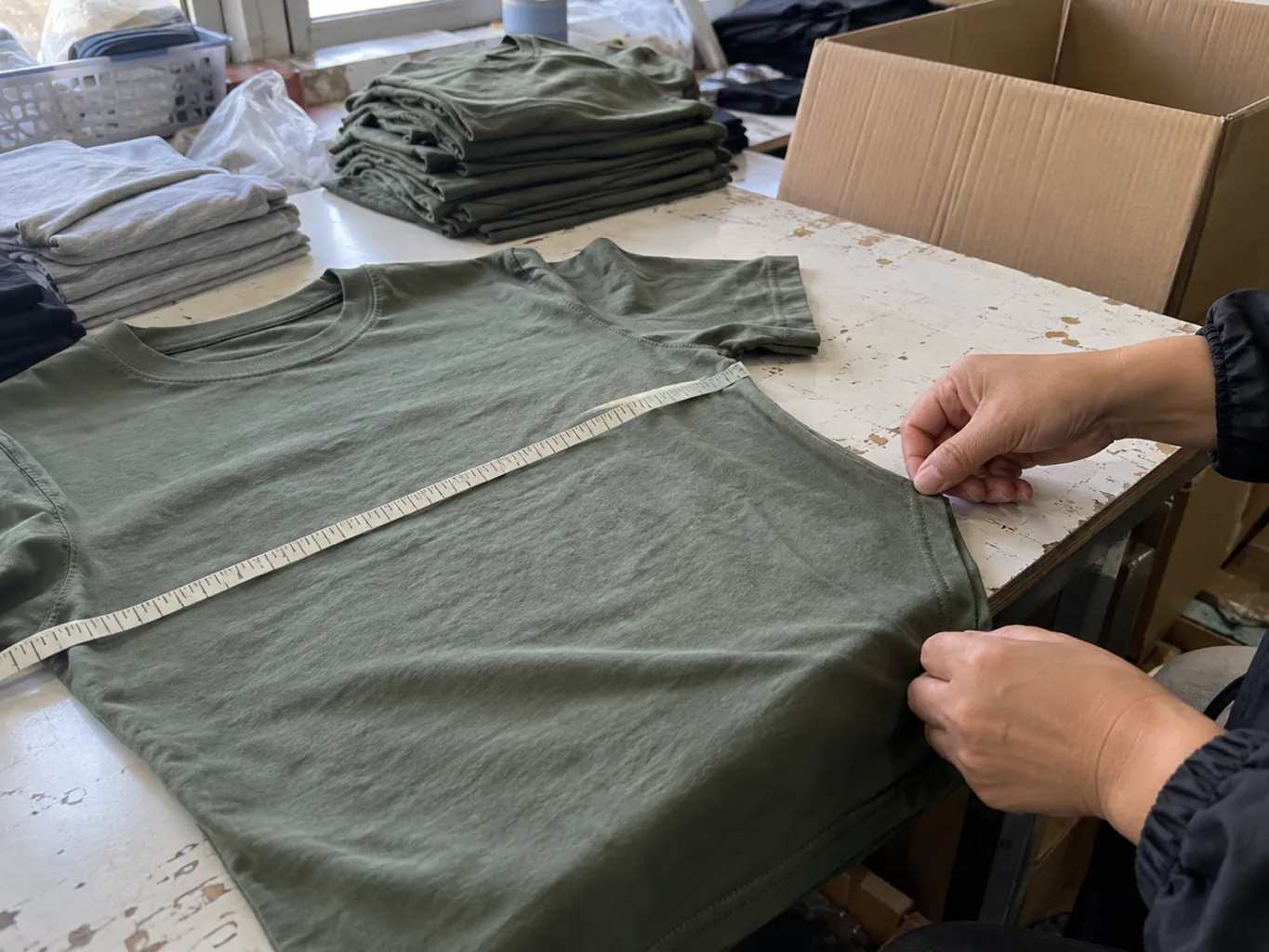 Hands checking a shirt seam beside a measuring tape and an open carton.