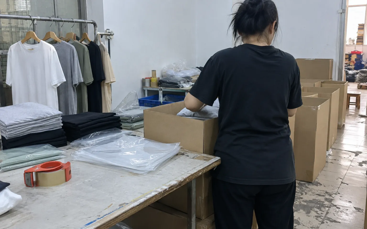 Worker packing folded garments into plain shipping cartons beside hanging clothing samples.