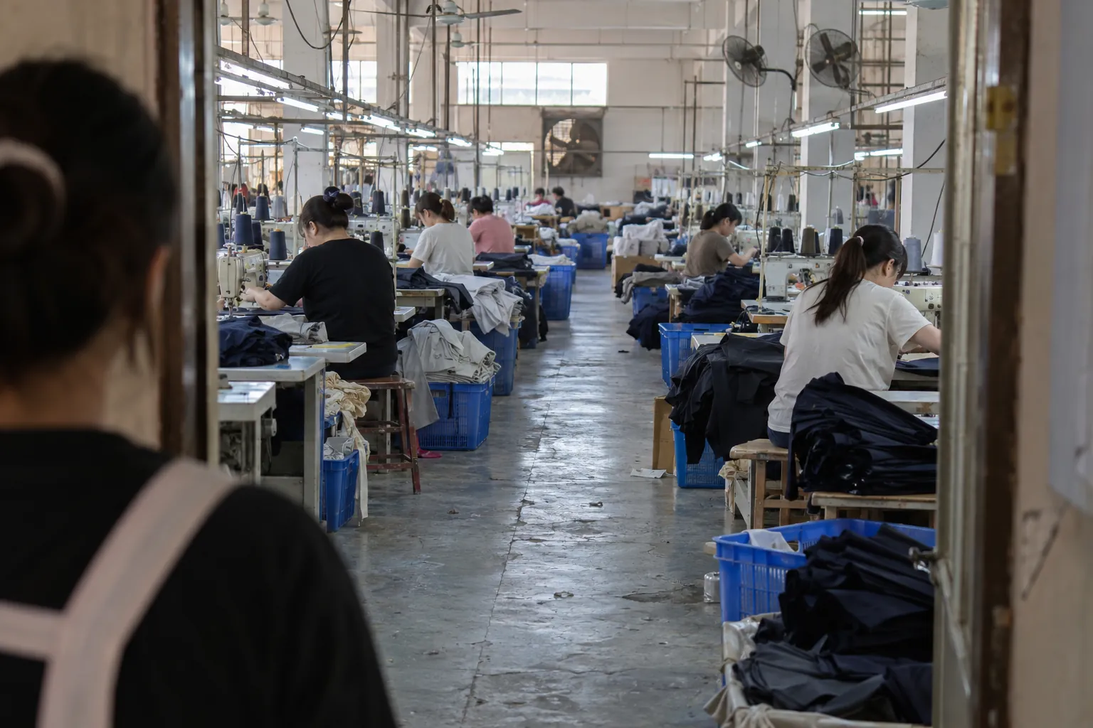 View past a person's shoulder into a sewing floor aisle of industrial machines and fabric bins